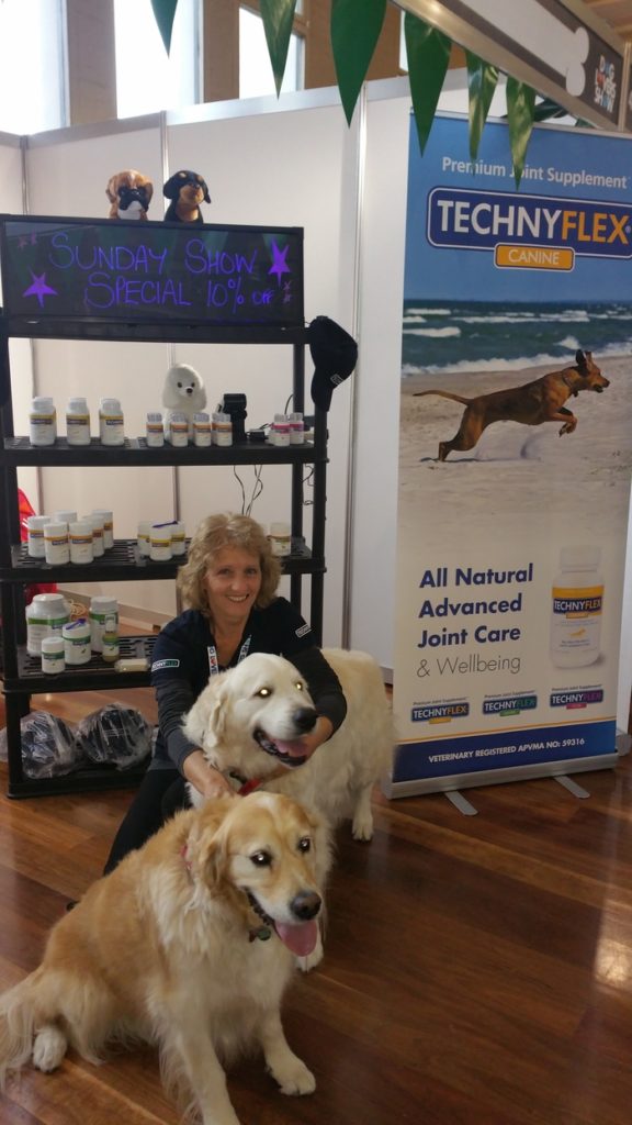 Liz Fields with two Golden Retrievers in front of a dog supplemens stand